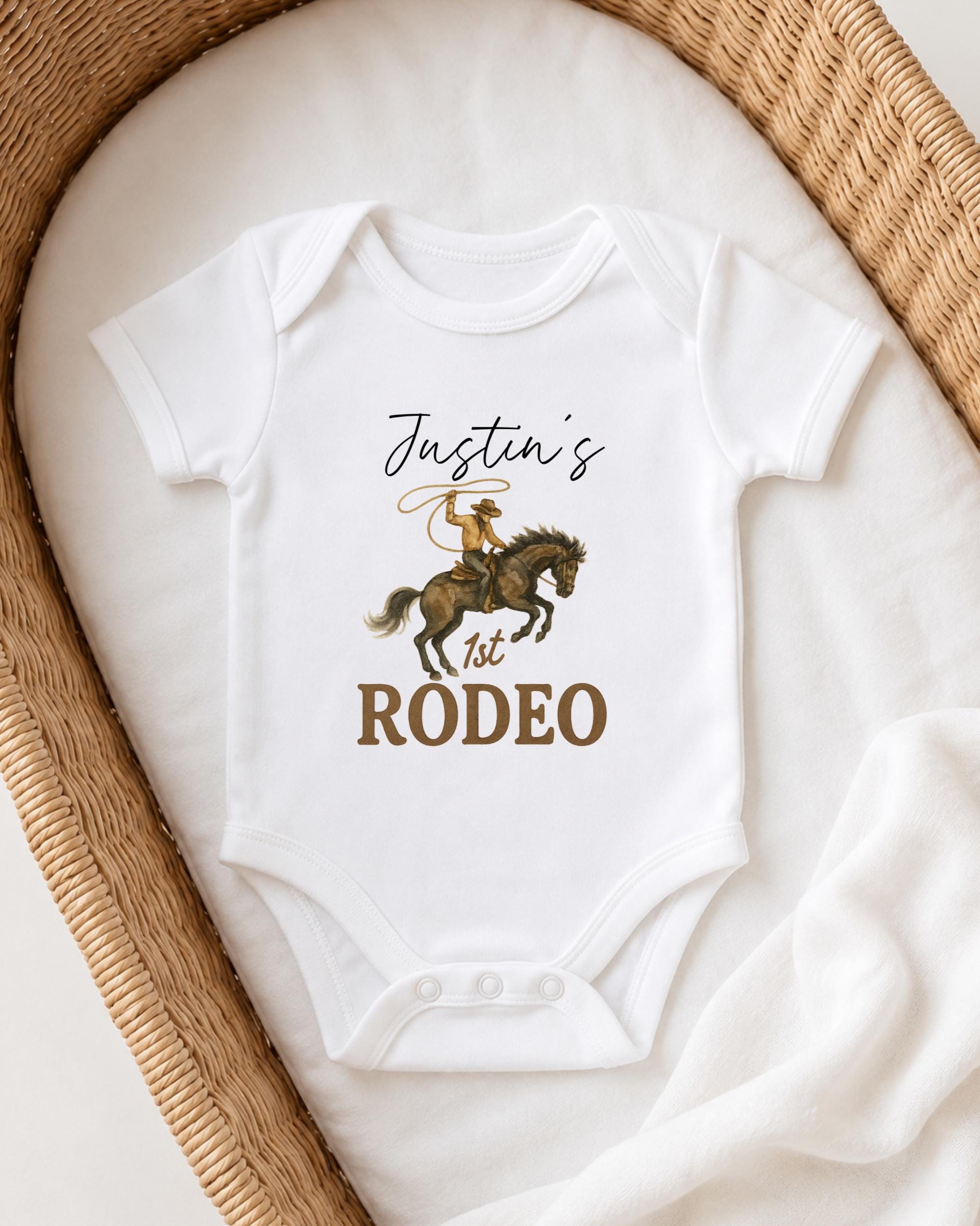 A white onesie with a cowboy graphic and the text " Justin's 1st Rodeo" printed on it, laid out on a white cloth.