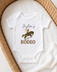 A white onesie with a cowboy graphic and the text " Justin's 1st Rodeo" printed on it, laid out on a white cloth.