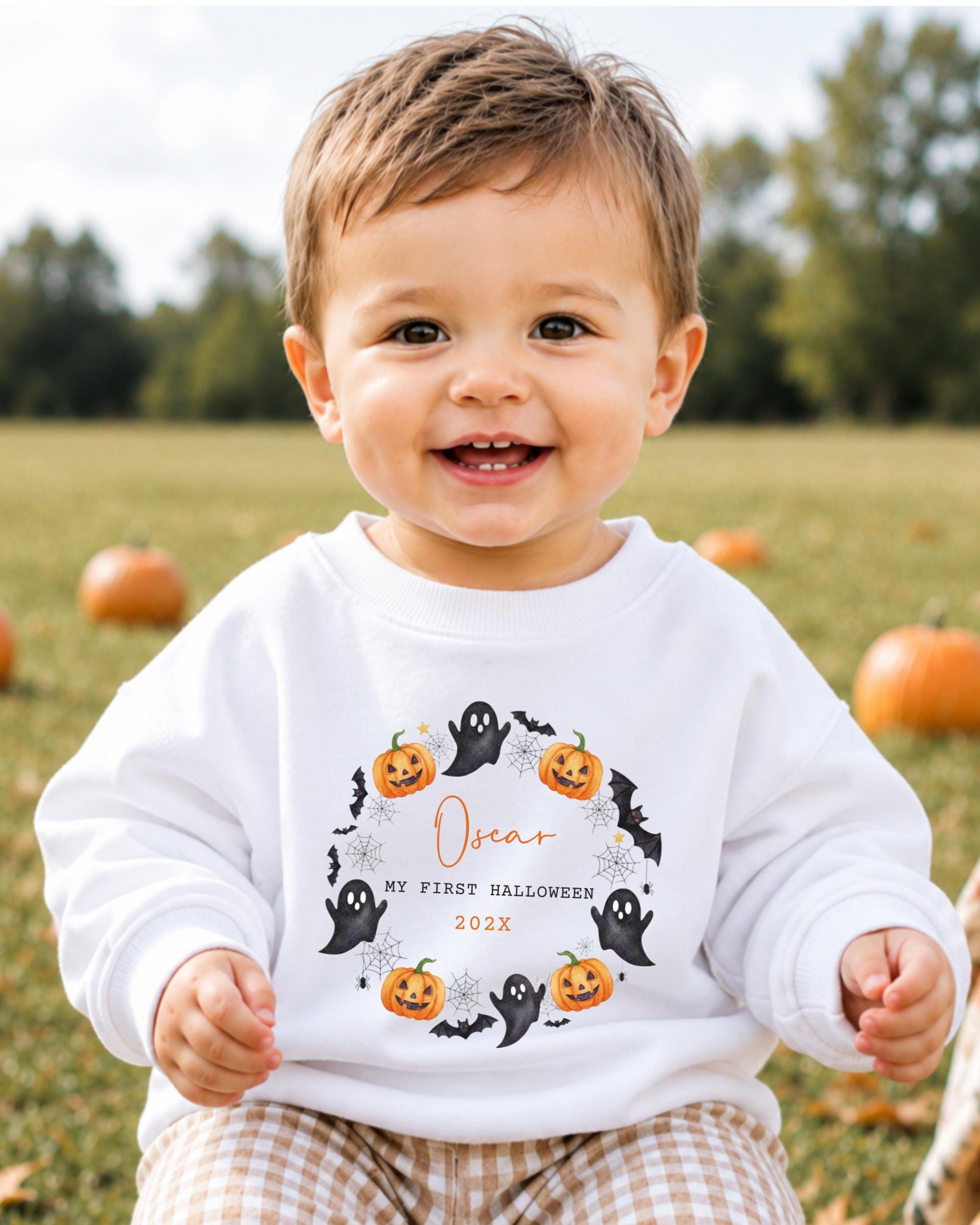 A smiling baby boy wearing a white sweatshirt with a Halloween-themed design, sitting in a field with pumpkins in the background.