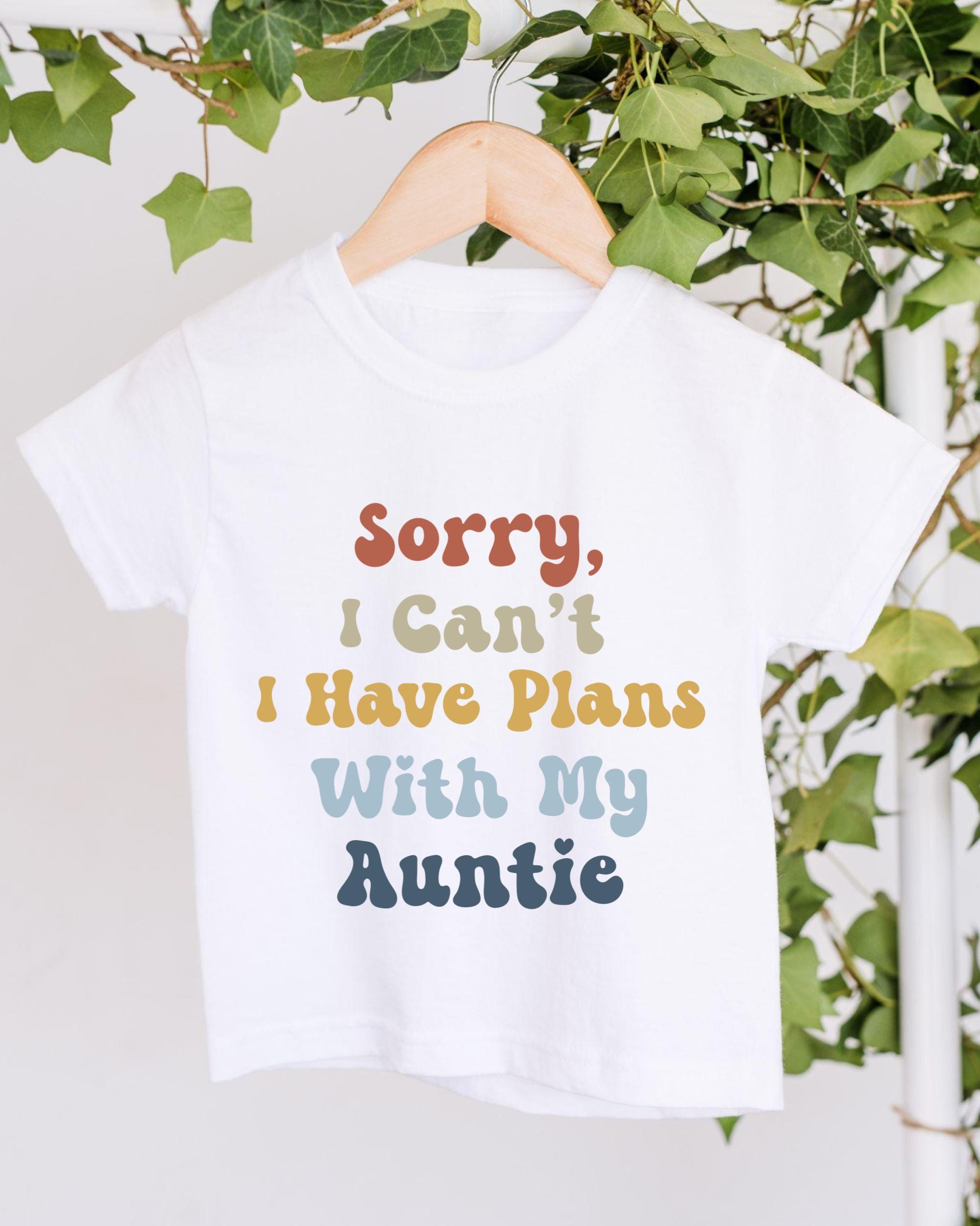 A white t-shirt with a colorful text design that says "Sorry, I Can't I Have Plans With My Auntie" hanging on a wooden hanger.