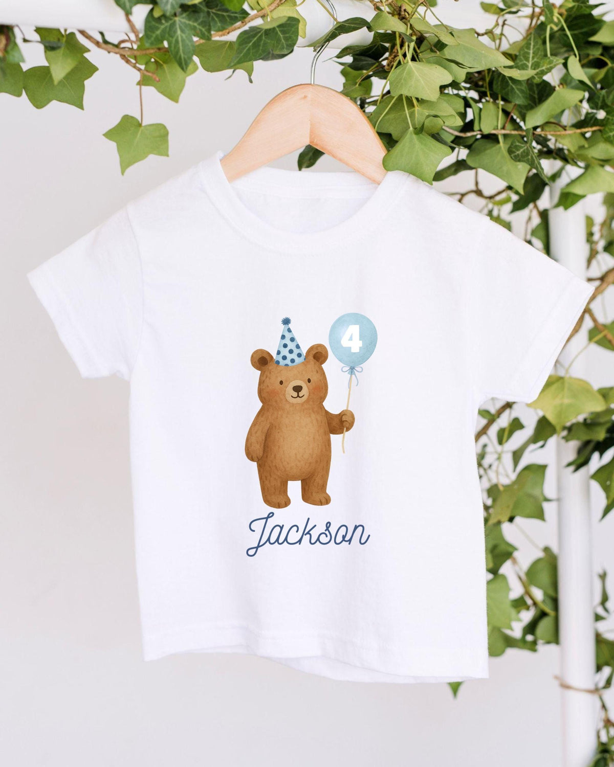 A white t-shirt with a cartoon image of a bear wearing a party hat and holding a blue balloon with the number 4 on it, along with the name "Jackson" written below the image.