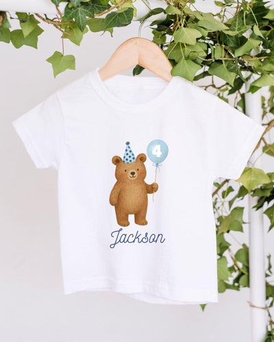 A white t-shirt with a cartoon image of a bear wearing a party hat and holding a blue balloon with the number 4 on it, along with the name "Jackson" written below the image.