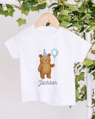 A white t-shirt with a cartoon image of a bear wearing a party hat and holding a blue balloon with the number 4 on it, along with the name "Jackson" written below the image.
