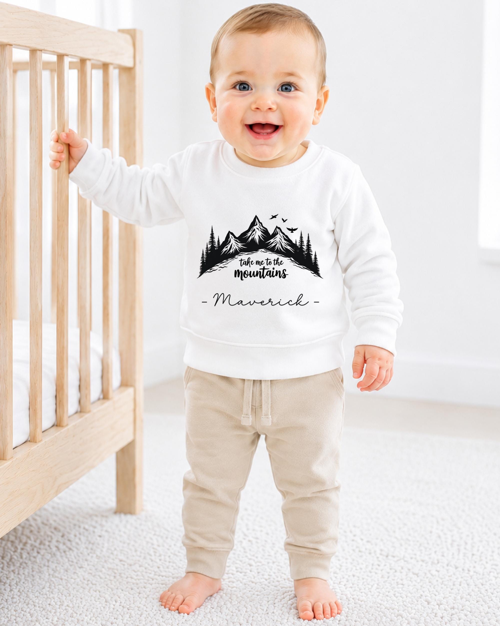 A smiling baby boy wearing a white sweatshirt with a mountain landscape design and the word "Maverick" printed on it, standing in a nursery setting.