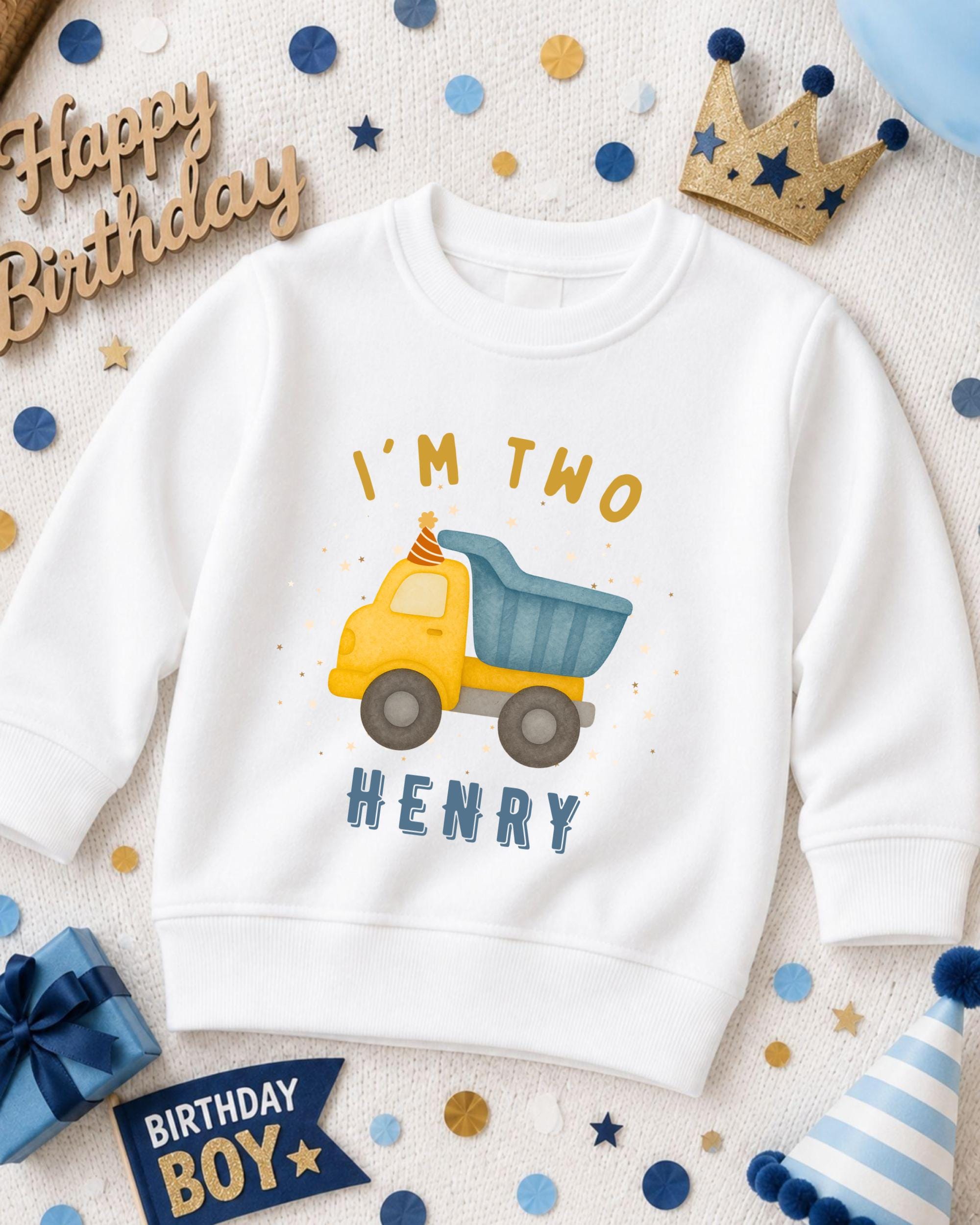 A white sweatshirt with a yellow truck graphic and the text "I'm two Henry" printed on it, surrounded by birthday decorations such as a crown, confetti, and a birthday sign.