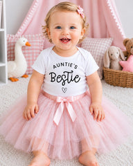 A smiling baby girl wearing a white t-shirt with the text "Auntie's Bestie" and a pink tutu skirt, sitting on a white carpet surrounded by stuffed animals.