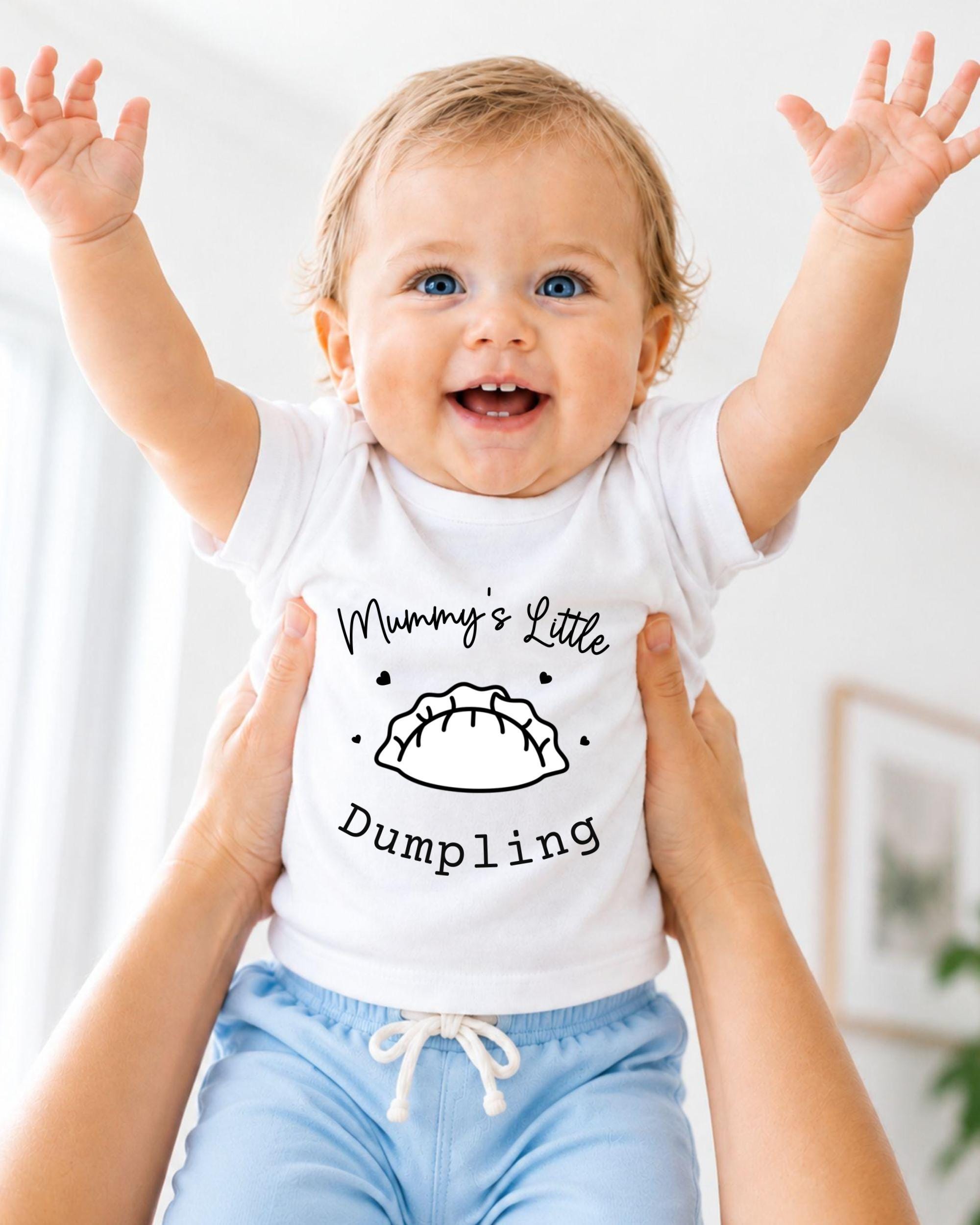 A smiling baby wearing a white t-shirt with a graphic and text printed on it, being held up by someone's arms.