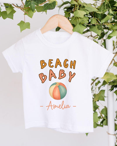 A white t-shirt with the text "BEACH BABY" and a beach ball graphic printed on it, hanging on a wooden hanger.