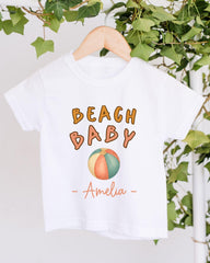 A white t-shirt with the text "BEACH BABY" and a beach ball graphic printed on it, hanging on a wooden hanger.