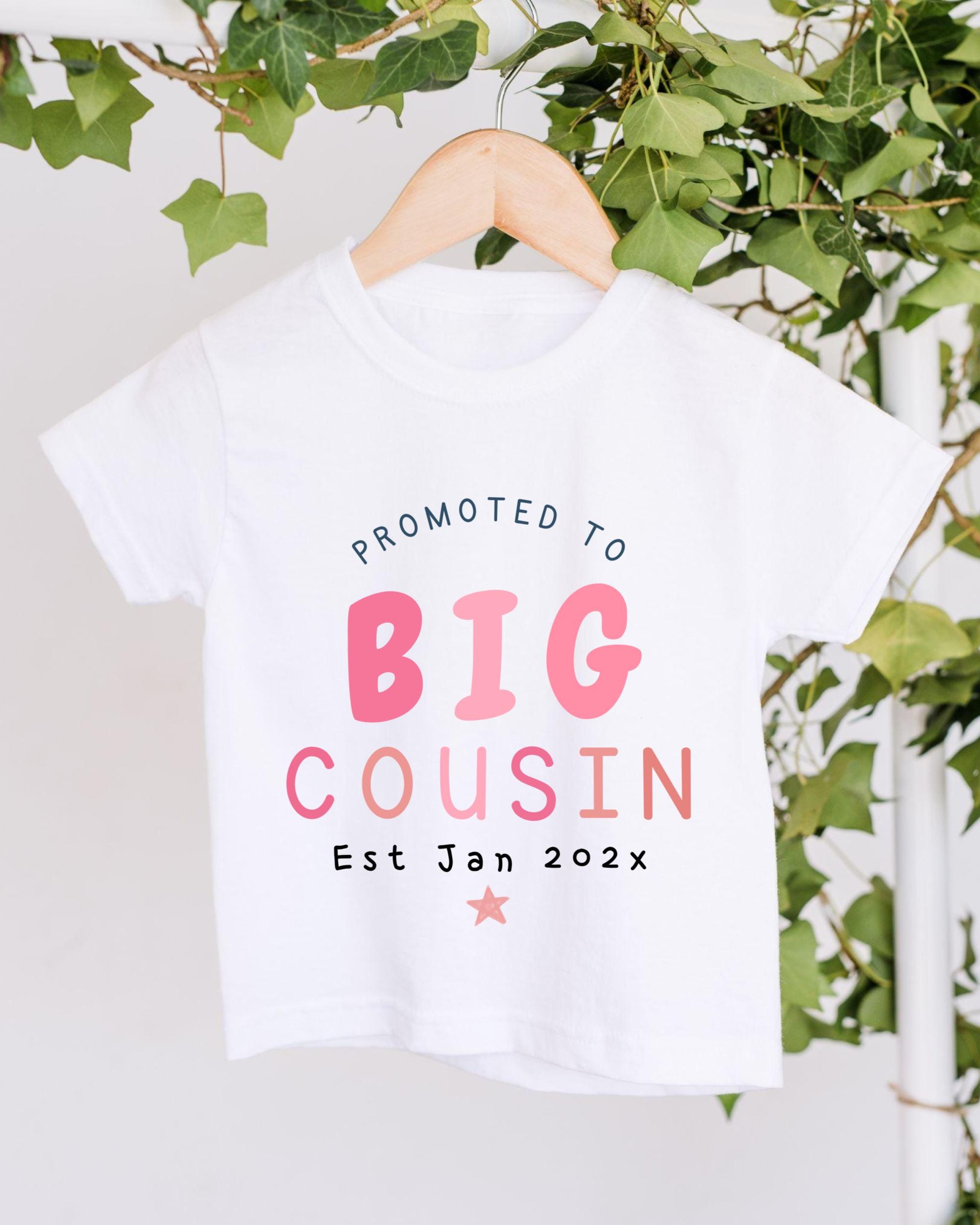 A white t-shirt with pink and blue text that reads "PROMOTED TO BIG COUSIN Est Jan 202x" hanging on a wooden hanger.