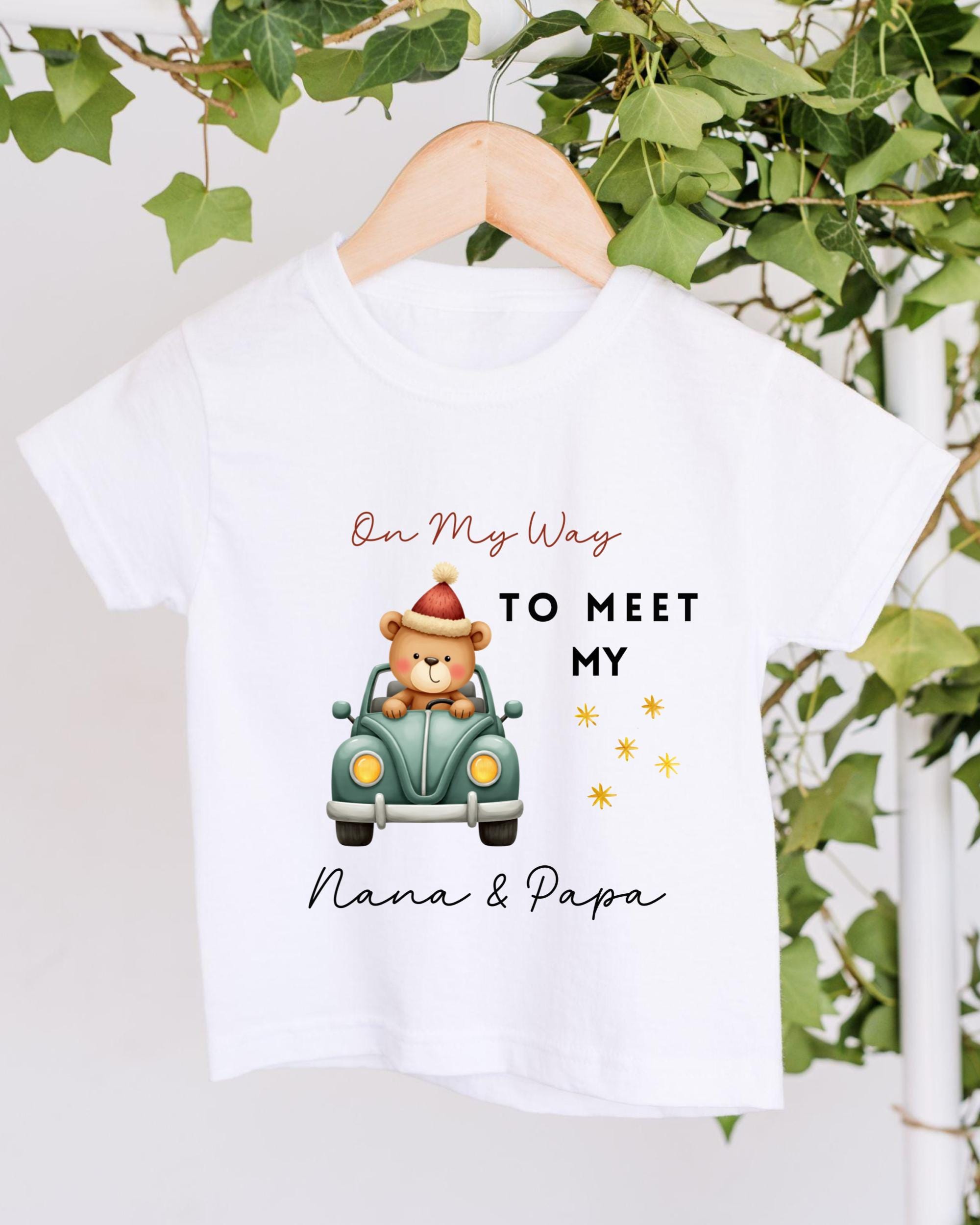 A white t-shirt with a graphic design featuring a teddy bear in a car, along with text that says "On My Way to Meet My Nana & Papa".