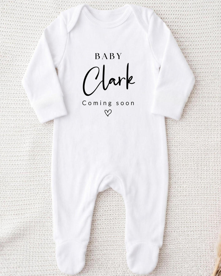 A white bodysuit with the text "BABY Clark Coming soon" printed on it.