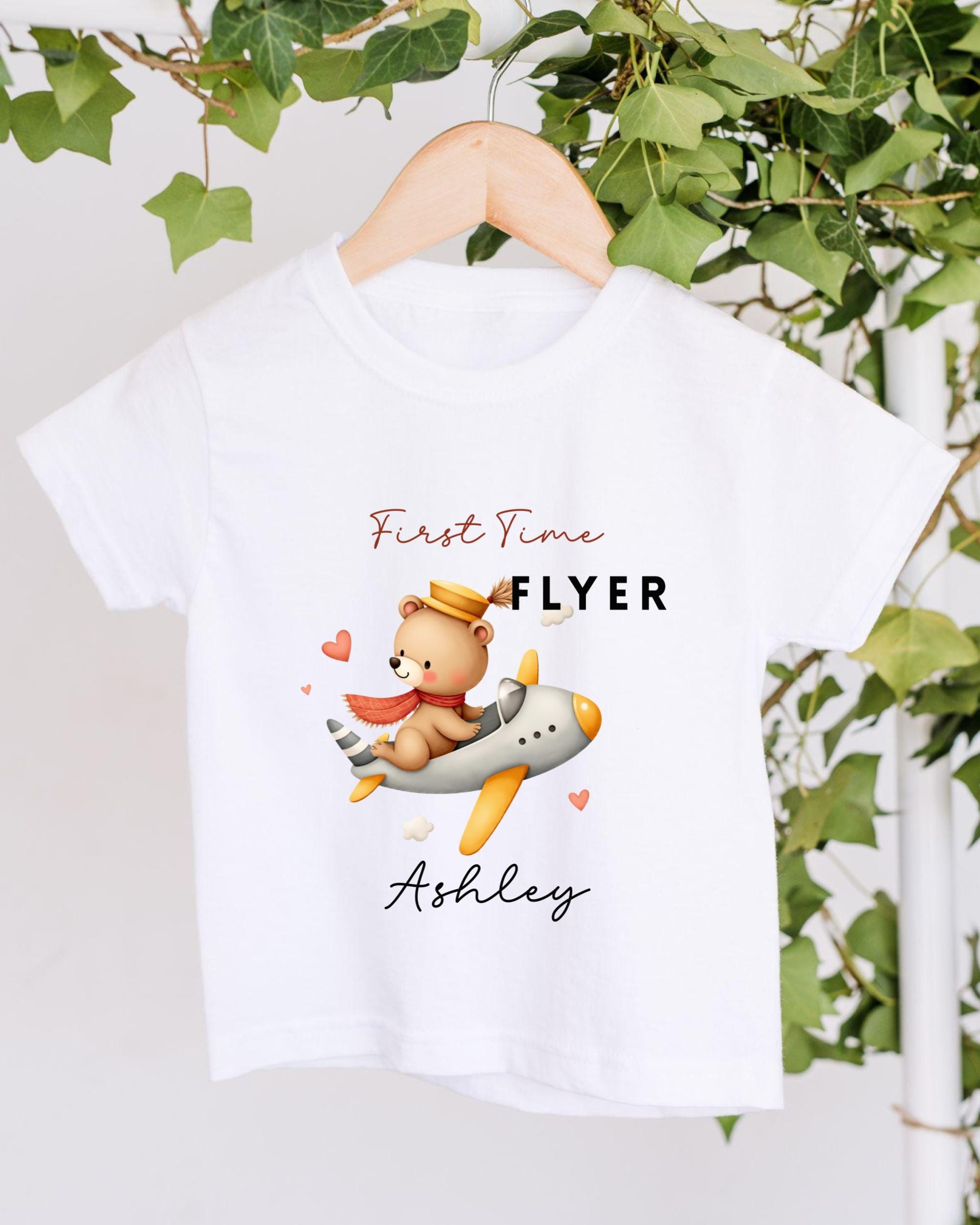 A white t-shirt with a cartoon image of a teddy bear flying on an airplane, along with the text "First Time Flyer Ashley" printed on it.