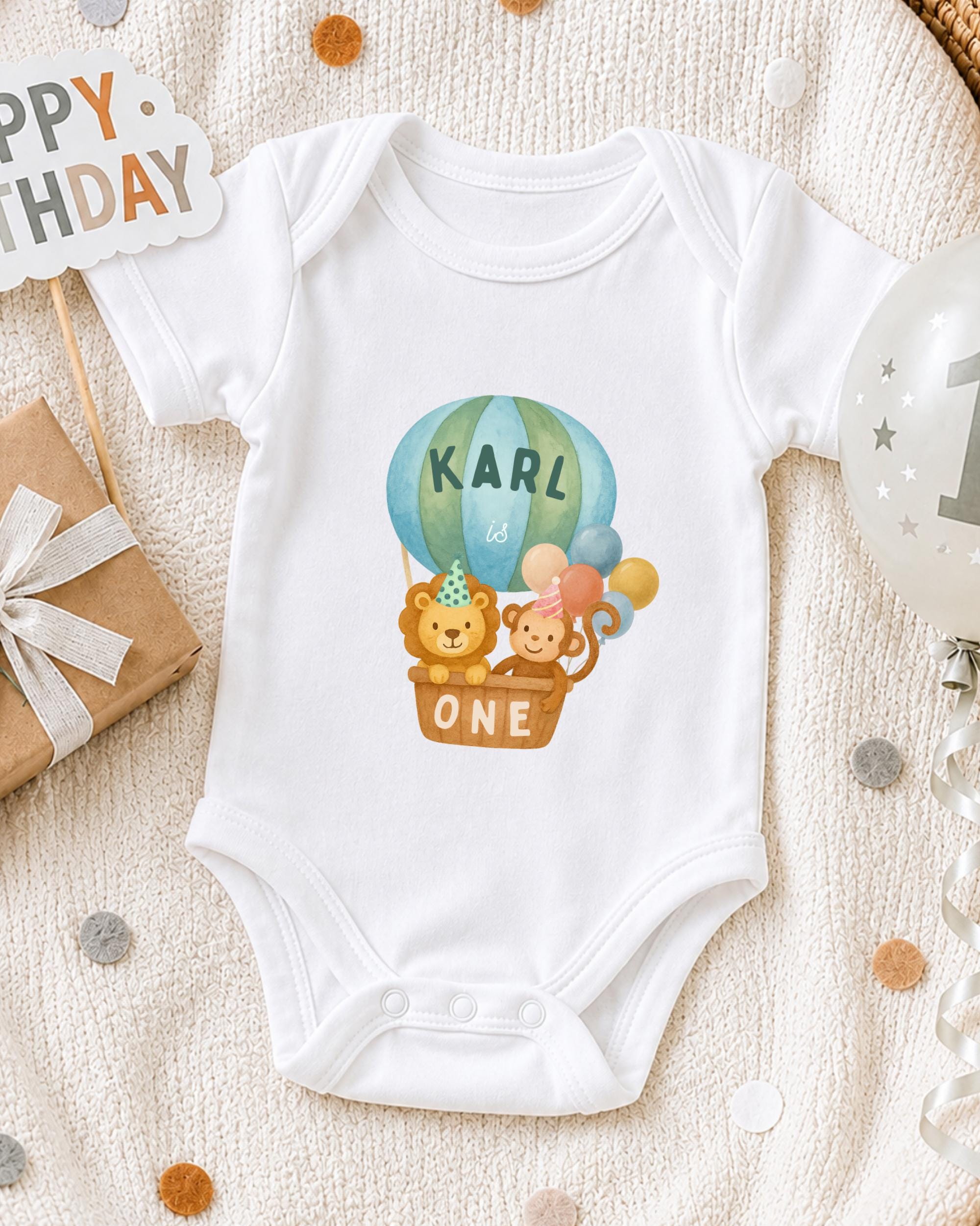A white onesie with a cartoon image of a monkey and a lion, along with the text "KARL is ONE" printed on it, is displayed on a bed with a "Happy Birthday" sign and a balloon.