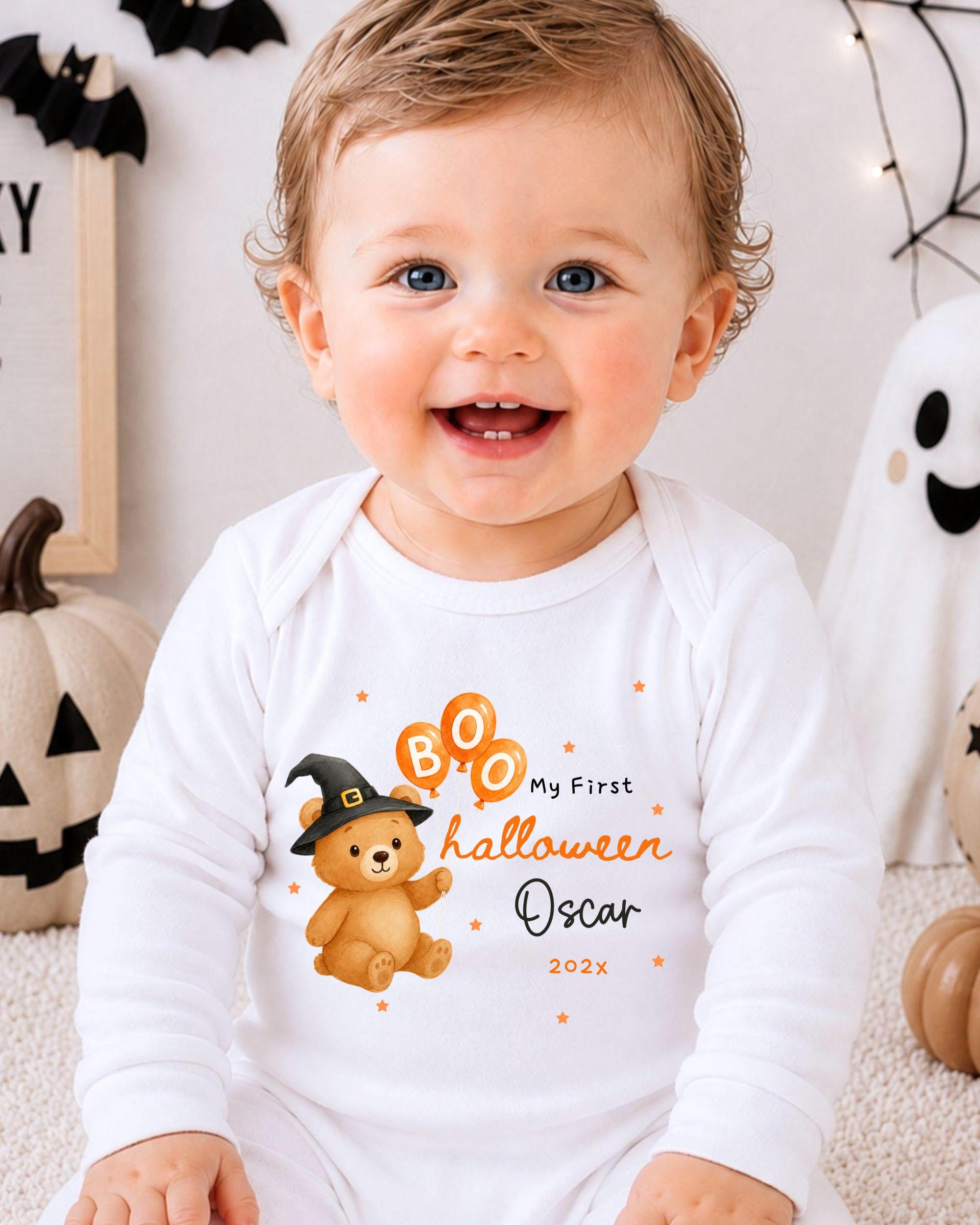 A smiling baby boy wearing a white onesie with a Halloween-themed design, including a teddy bear and a witch's hat.