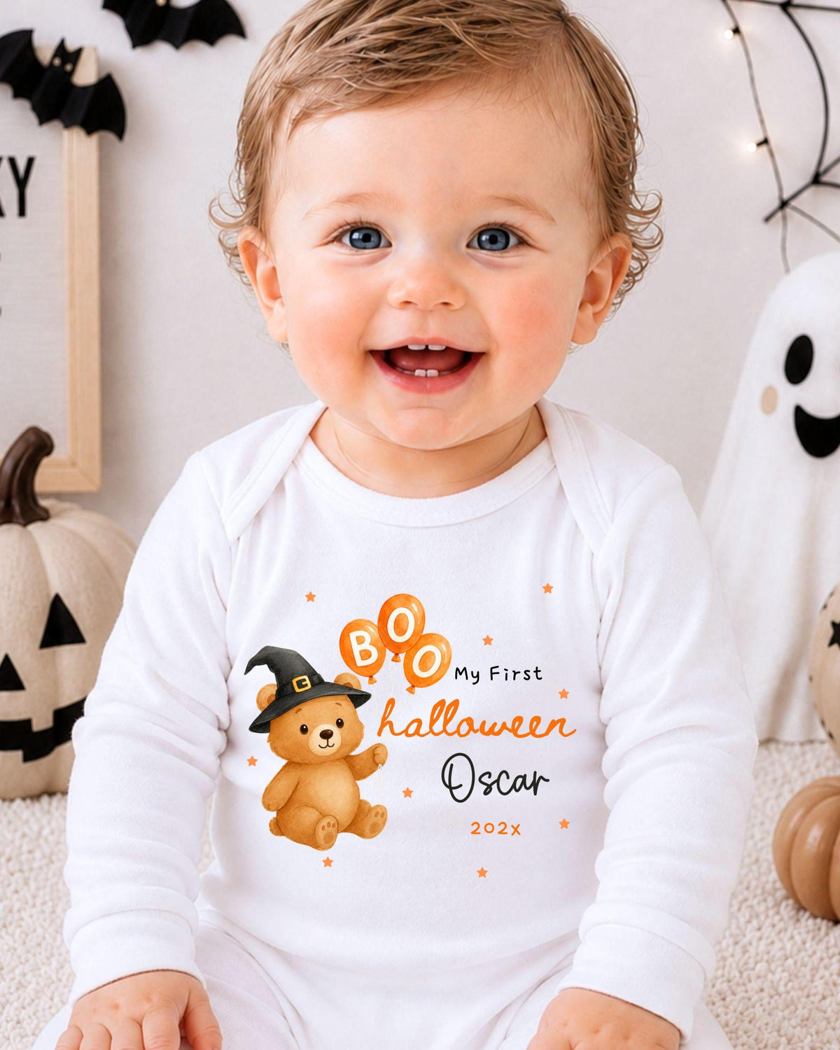 A smiling baby boy wearing a white onesie with a Halloween-themed design, including a teddy bear and a witch's hat.