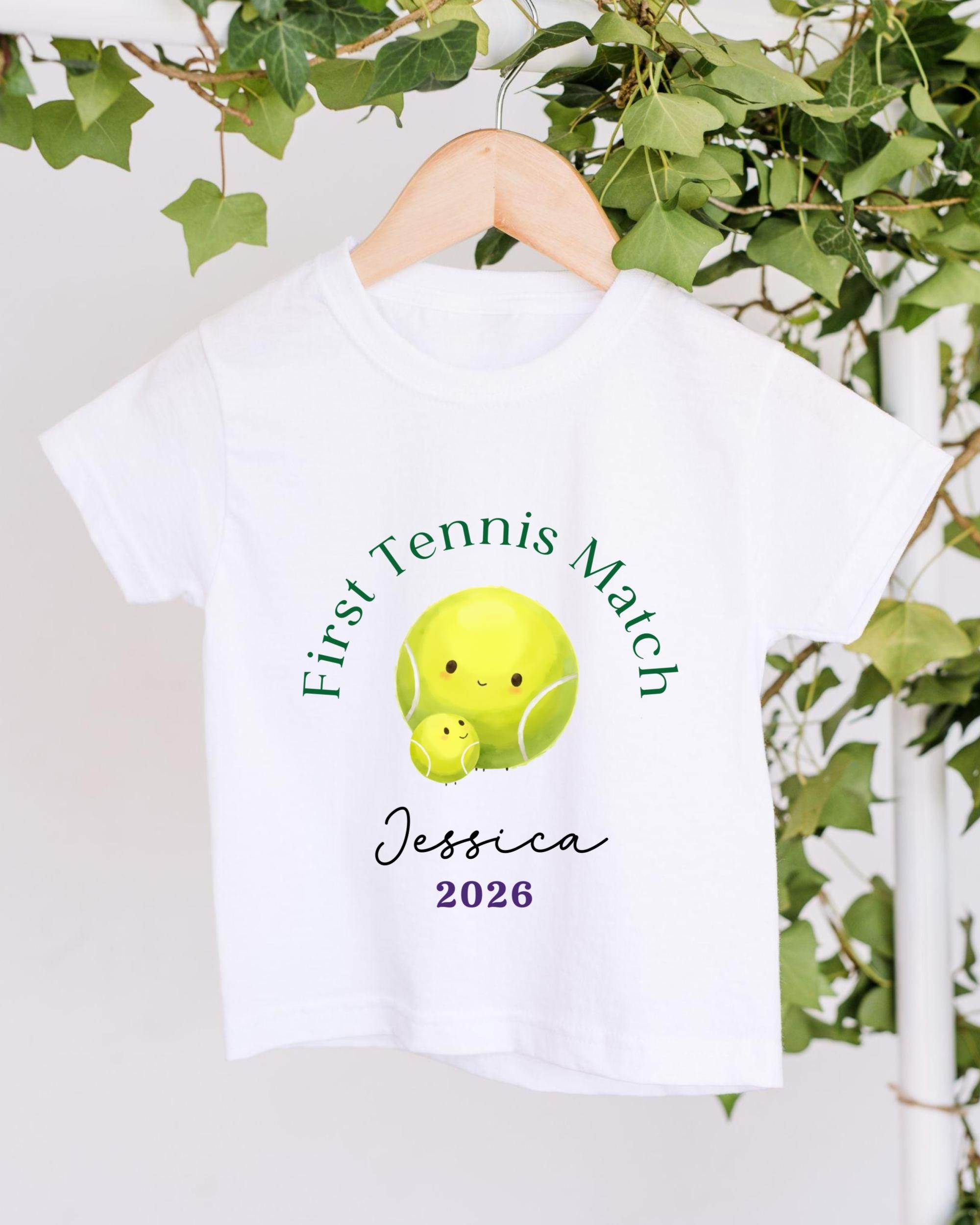 A white t-shirt with a tennis ball design and text printed on it, hanging on a wooden hanger.