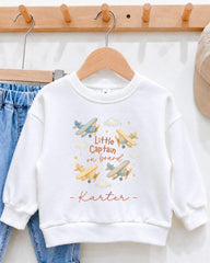 A white sweatshirt with a printed design of airplanes and clouds, along with the text "Little Captain on board - Kartter".
