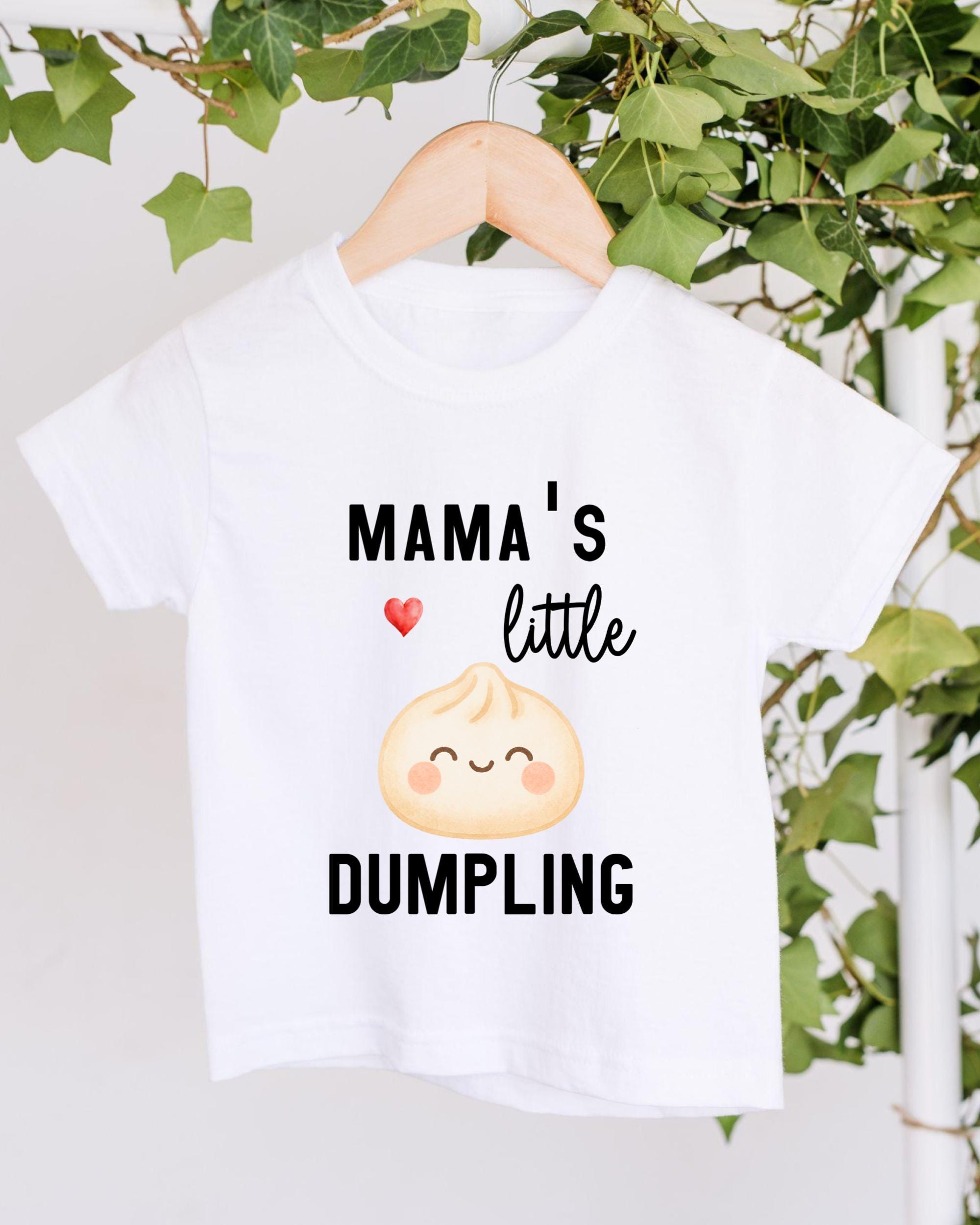 A white t-shirt with a printed design featuring a smiling dumpling character and the text "Mama's little DUMPLING".
