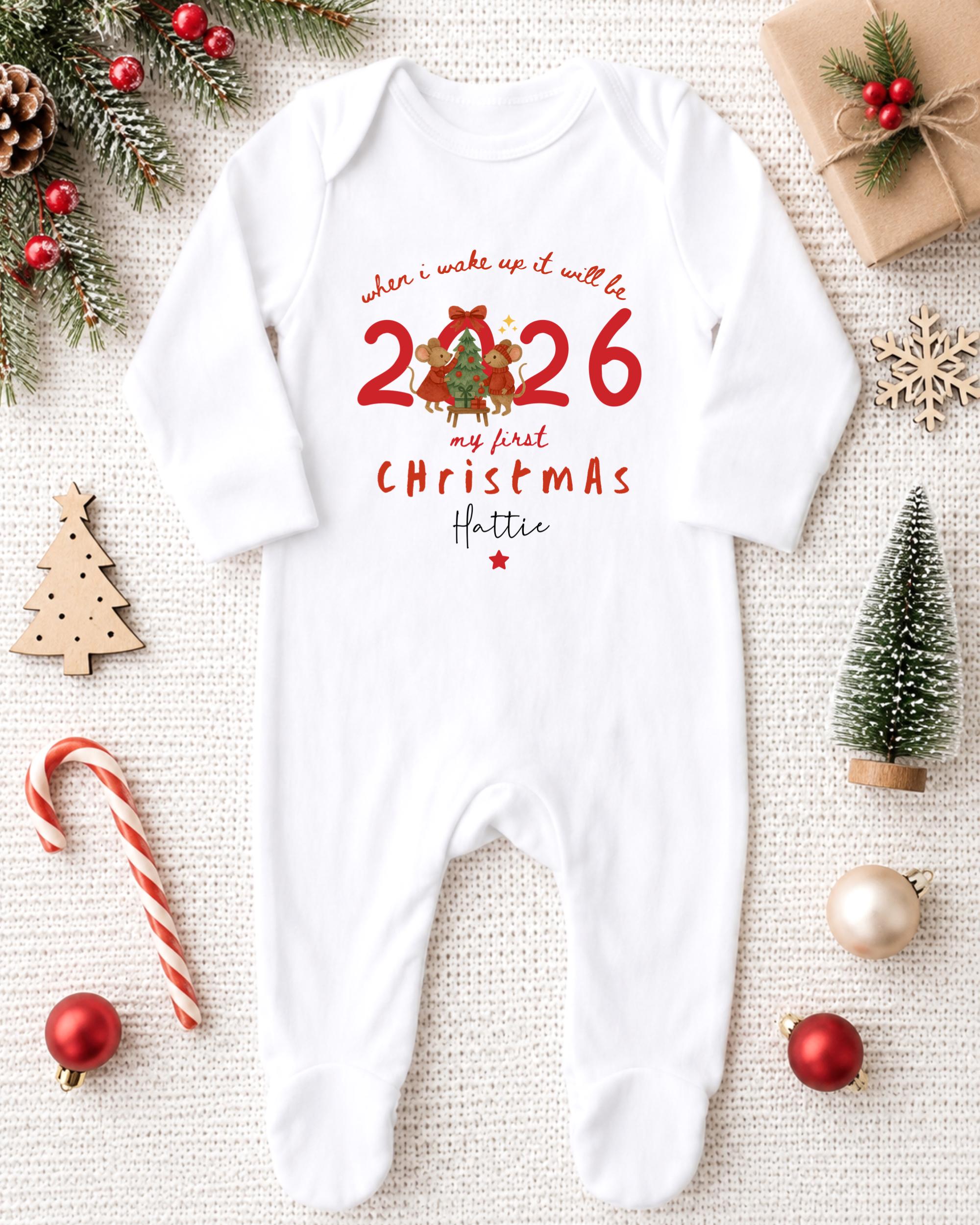 A white onesie with a Christmas-themed design, surrounded by holiday decorations such as a pine tree, ornaments, and a small Christmas tree.