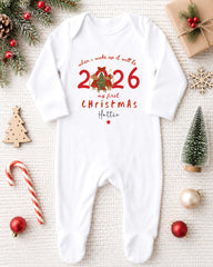 A white onesie with a Christmas-themed design, surrounded by holiday decorations such as a pine tree, ornaments, and a small Christmas tree.