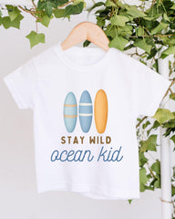 A white t-shirt with a graphic design featuring three surfboards and the text "STAY WILD ocean kid" printed on it.