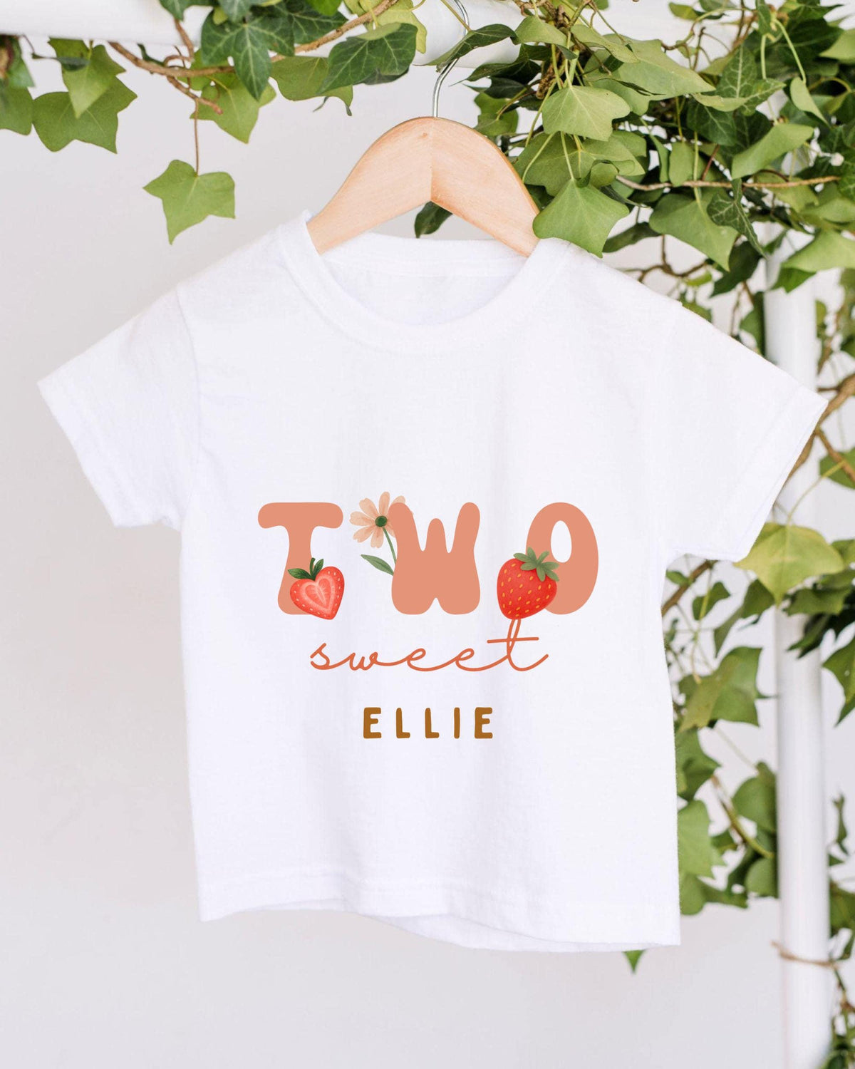 A white t-shirt with the text "TWO sweet Ellie" printed on it, along with illustrations of strawberries and leaves.