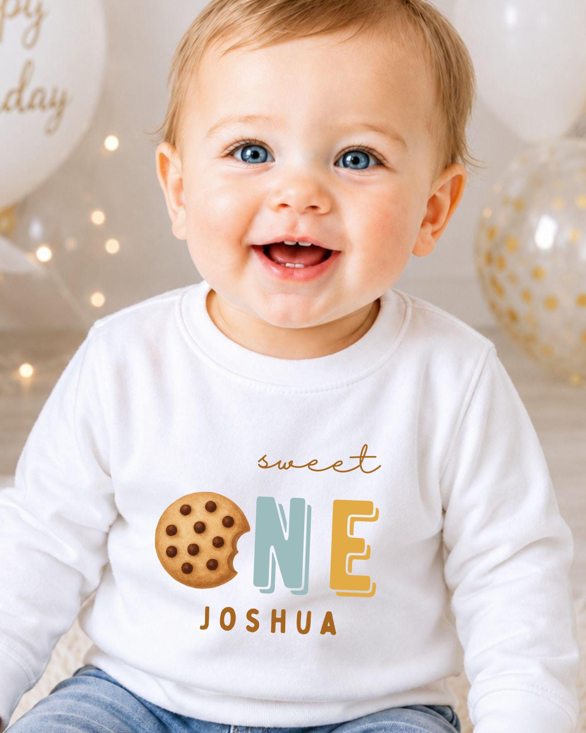A smiling baby boy wearing a white shirt with a cookie and the text "Sweet One Joshua" printed on it.