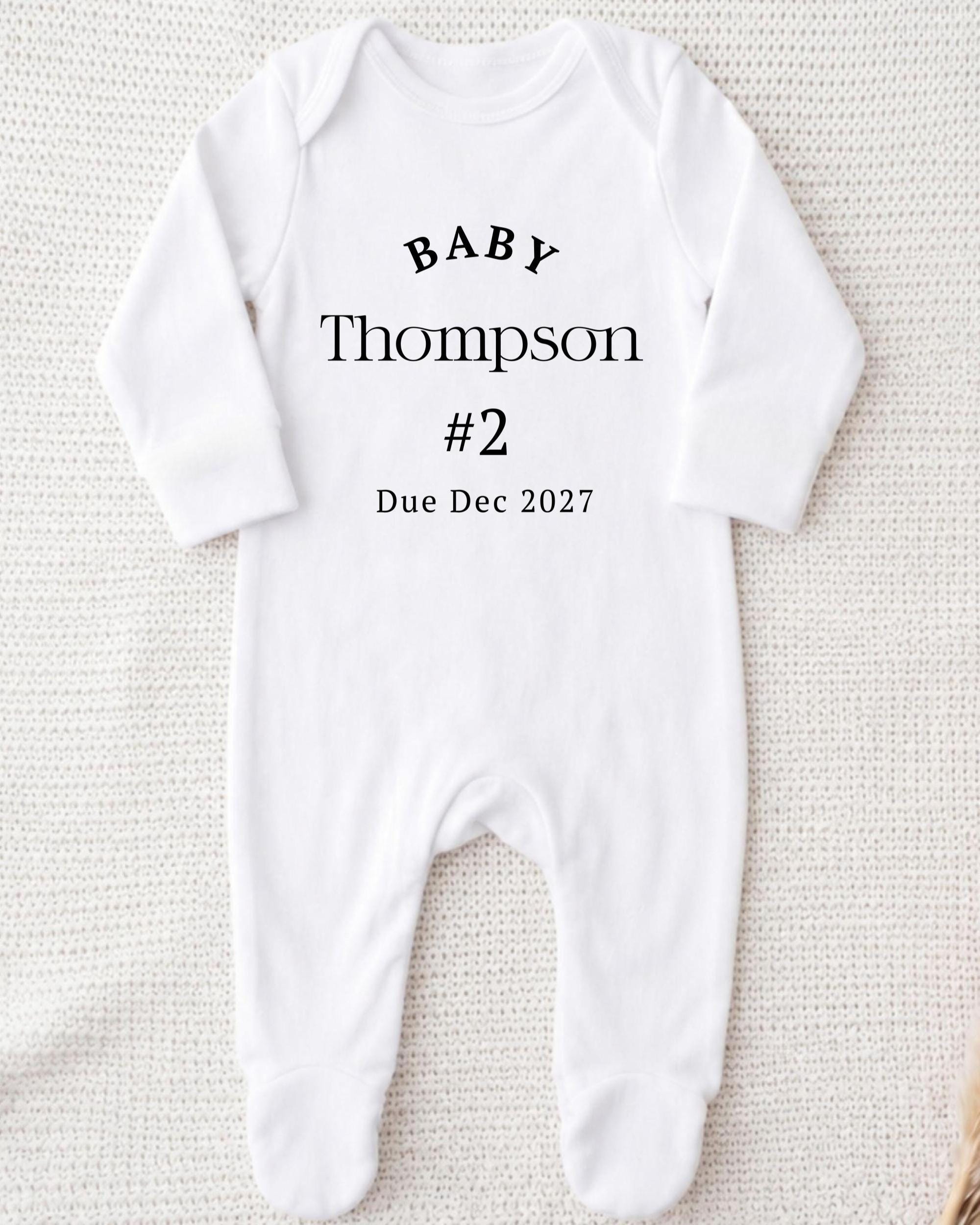 A white onesie with text printed on it, indicating the baby's name and the date of the baby's announcement.
