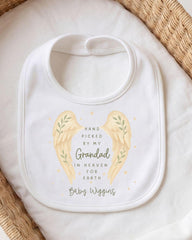 A white baby bib with a design featuring two angel wings and the text "Hand picked by my Grandfather in Heaven for Earth Baby Wiggins".