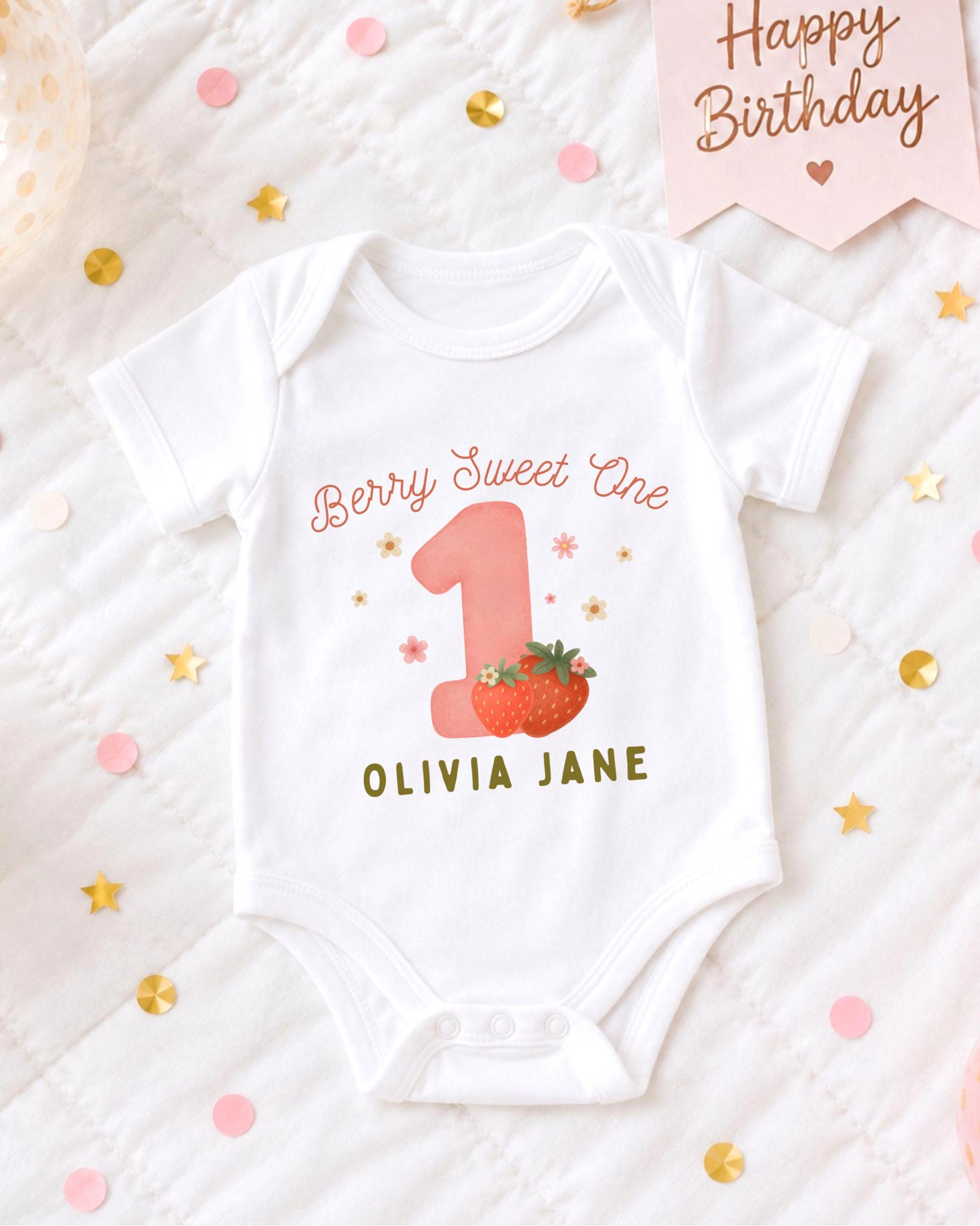 A white onesie with a pink number one and a strawberry graphic, along with the text "Berry Sweet One" and "OLIVIA JANE". The onesie is placed on a bed with confetti and a birthday card.