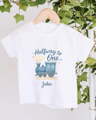 A white t-shirt with a graphic design of a blue train on a track, surrounded by stars and text that reads "Halfway to One John".