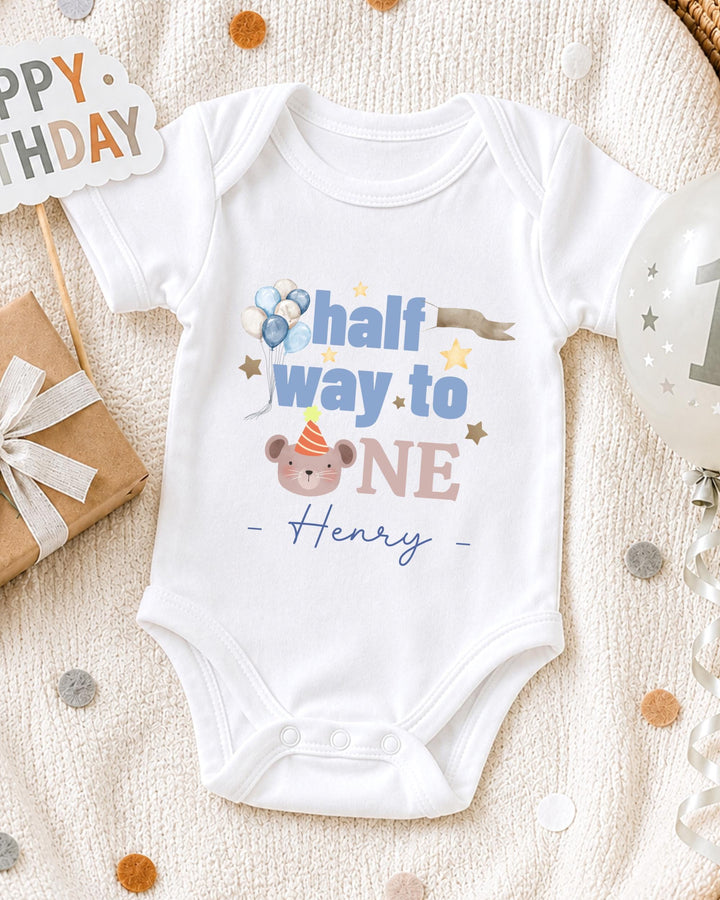 A white onesie with a personalized message, "Half way to one Henry", is displayed on a bed with a gift box and a birthday balloon.