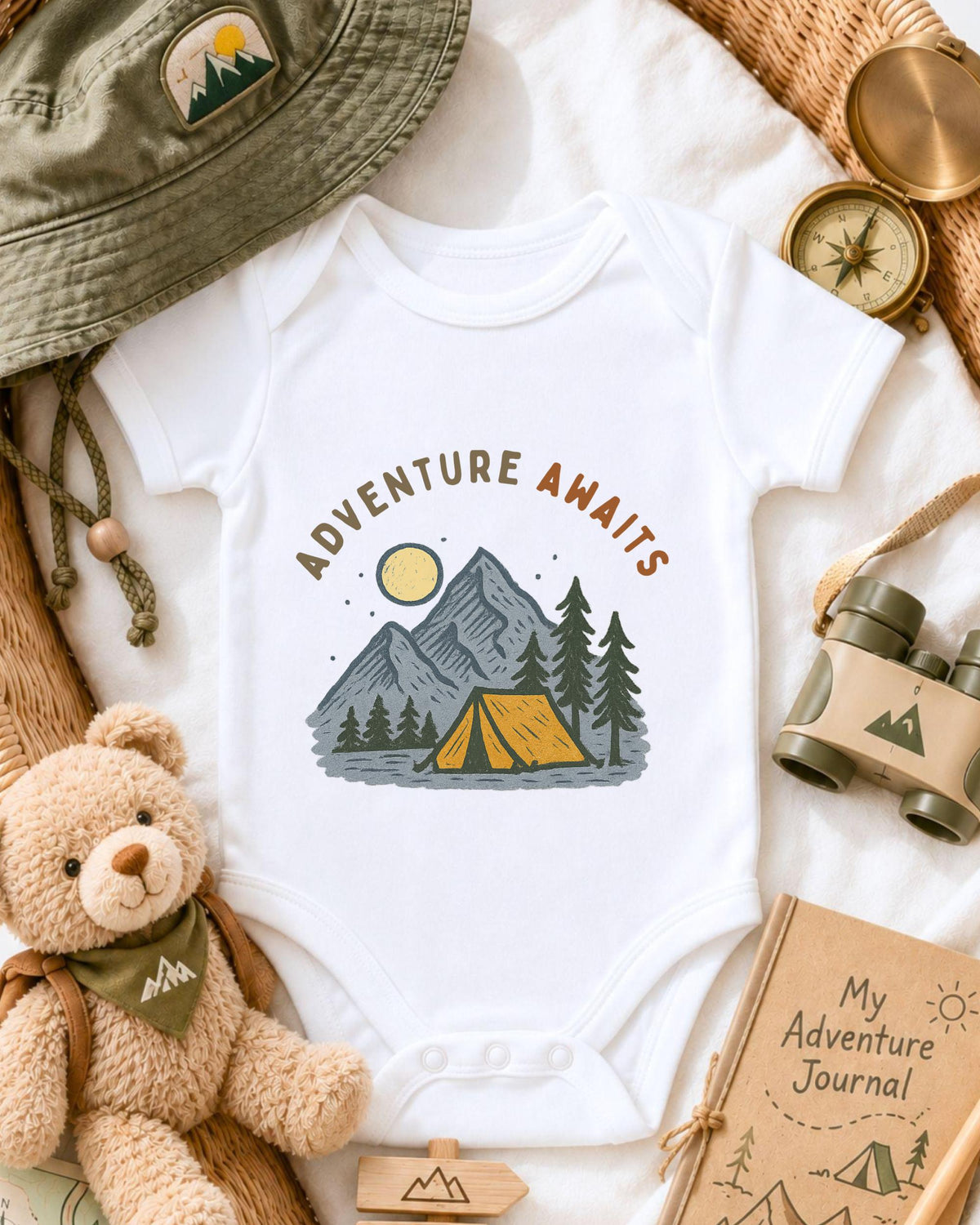 a baby onesie with a camping scene printed on it, surrounded by various camping-related items such as a hat, compass, binoculars, and a teddy bear.