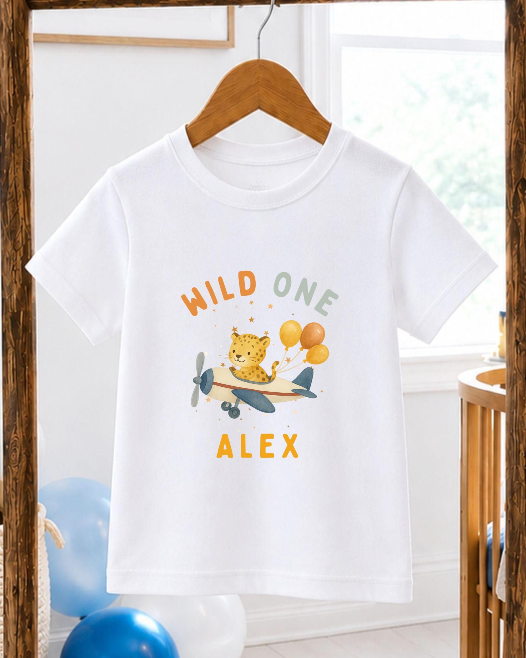 A white t-shirt with a printed design of a leopard and balloons, along with the text "WILD ONE ALEX" printed on it.