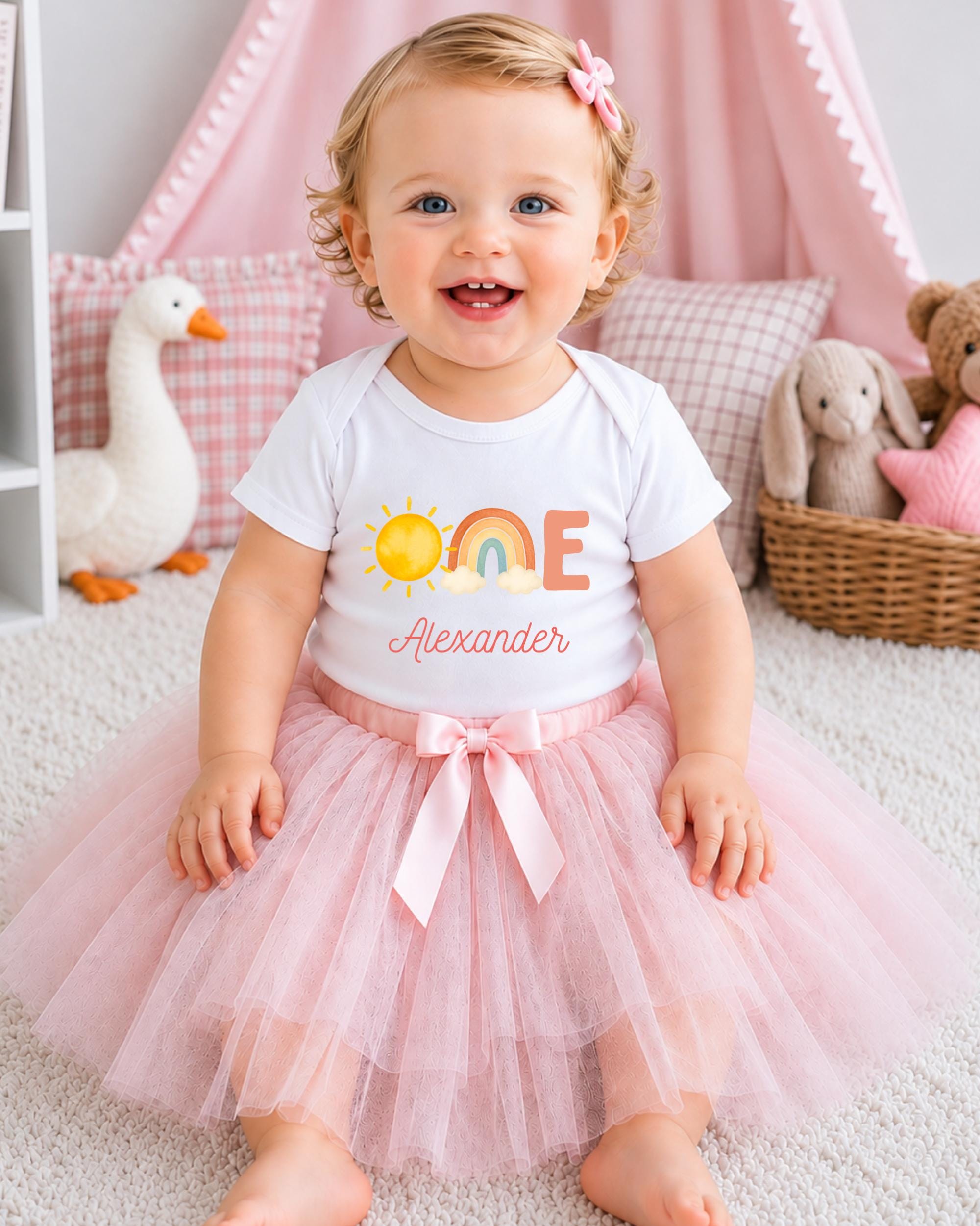 A smiling baby girl wearing a white t-shirt with a rainbow graphic and the name "Alexander" on it, sitting on a pink carpet surrounded by stuffed animals.