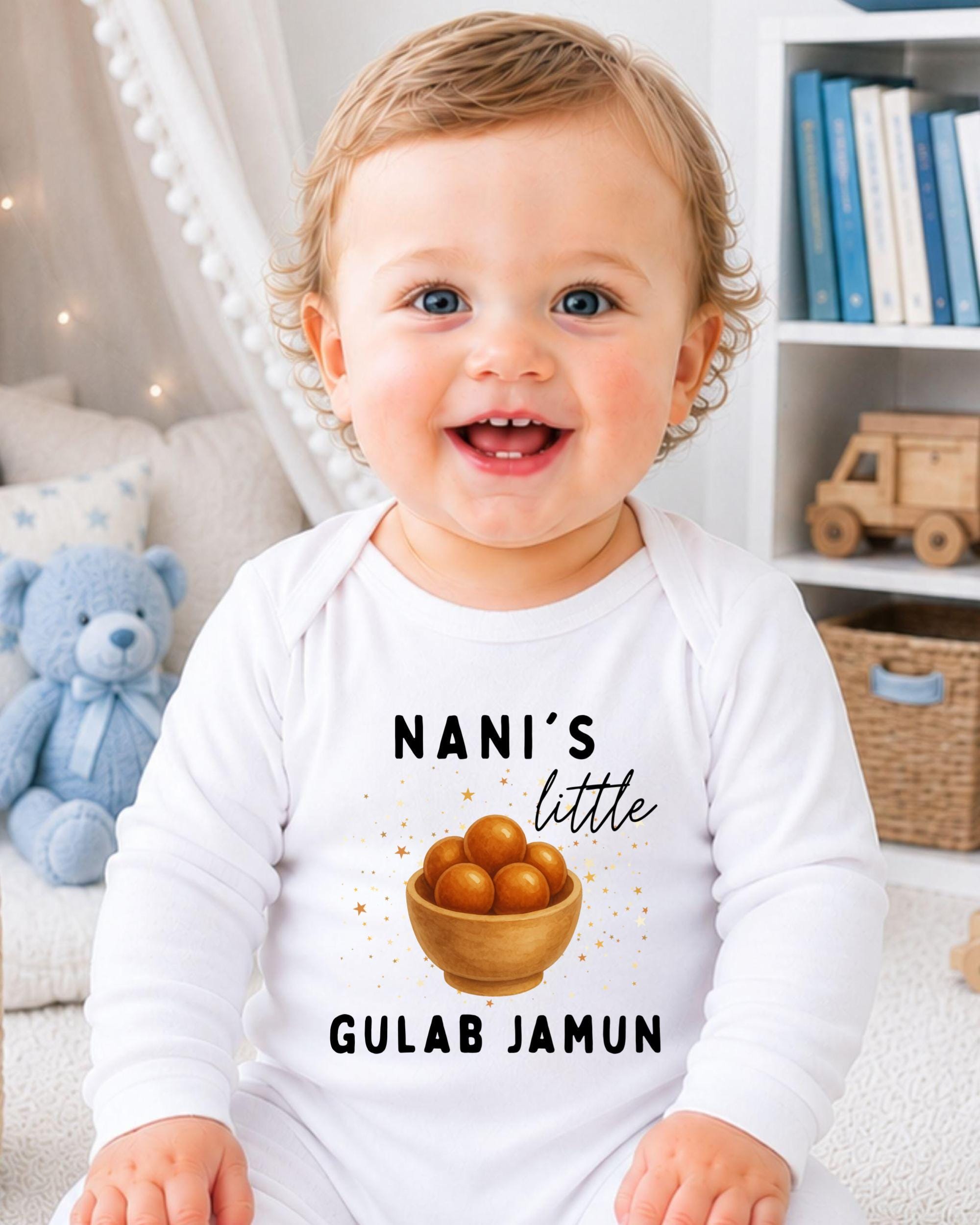 A smiling baby wearing a white onesie with a graphic and text printed on it, sitting in a room with a teddy bear and bookshelf in the background.