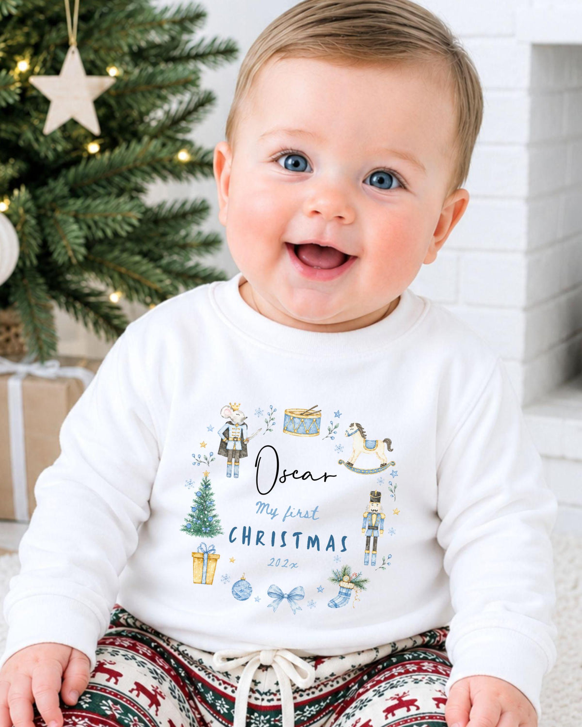 A smiling baby boy wearing a white sweatshirt with a personalized design and text, sitting in front of a Christmas tree.