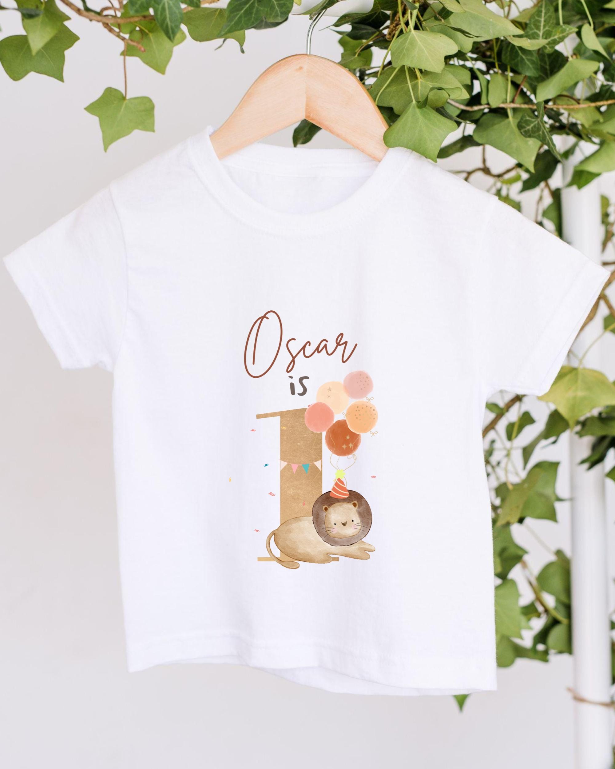 A white t-shirt with a graphic design featuring a lion and balloons, along with the text "Oscar is" printed on it.