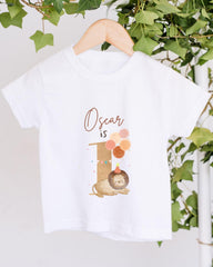 A white t-shirt with a graphic design featuring a lion and balloons, along with the text "Oscar is" printed on it.