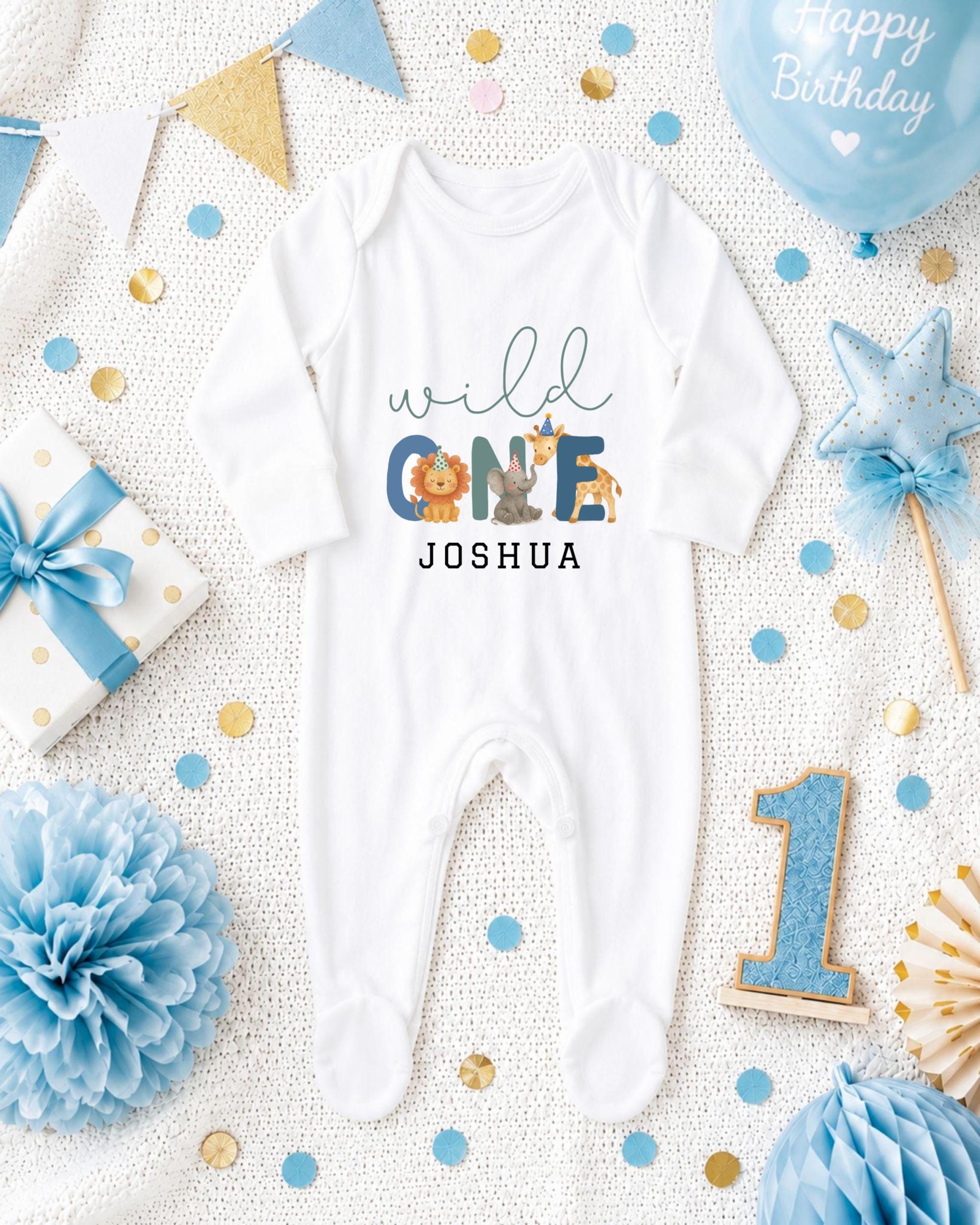 A white onesie with the text "wild one Joshua" printed on it, surrounded by various baby-themed items such as a blue balloon, a blue and gold star, and a blue and gold bow.