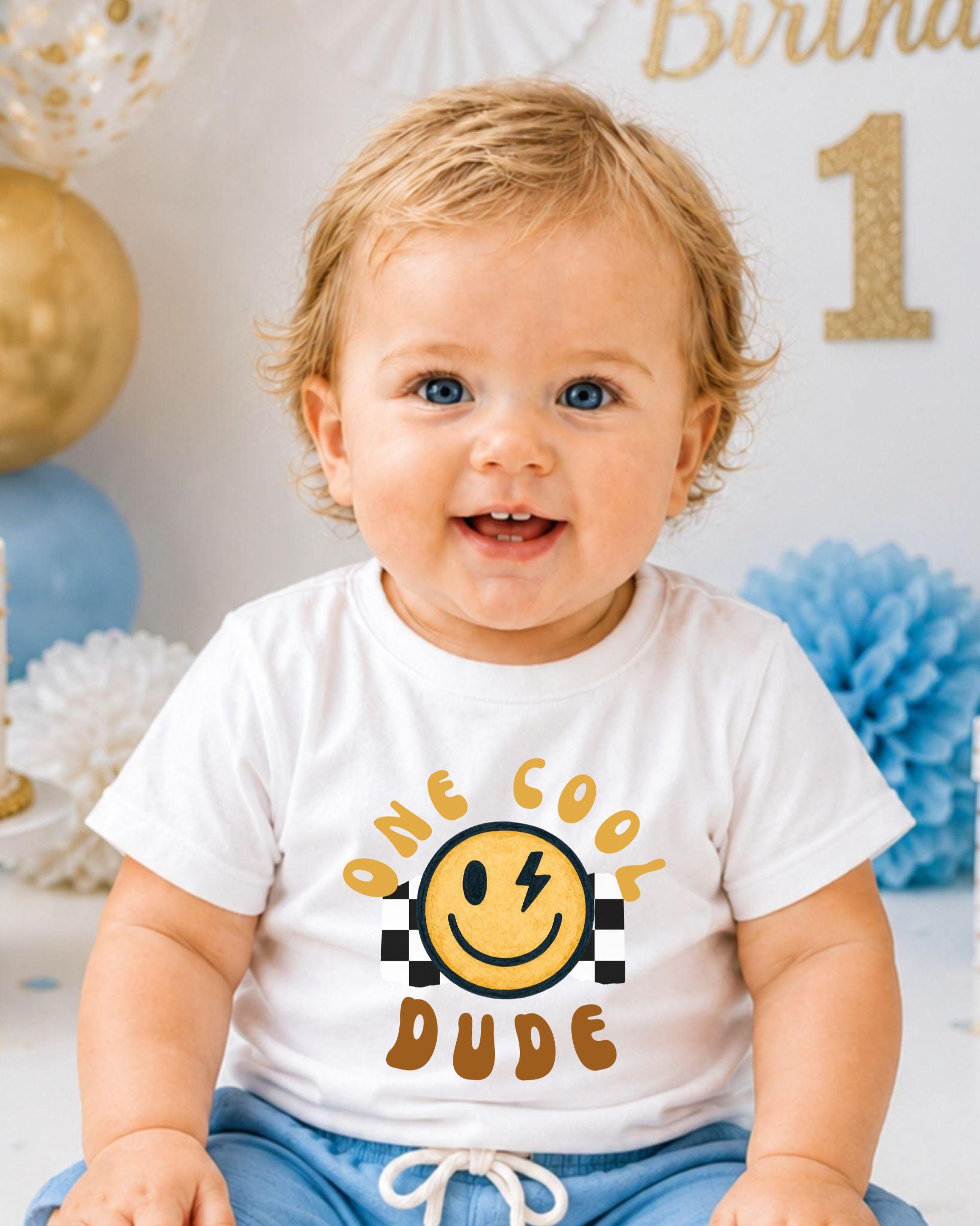 A smiling baby boy wearing a white t-shirt with a smiley face and the text "One cool dude" is sitting in front of a birthday party backdrop.