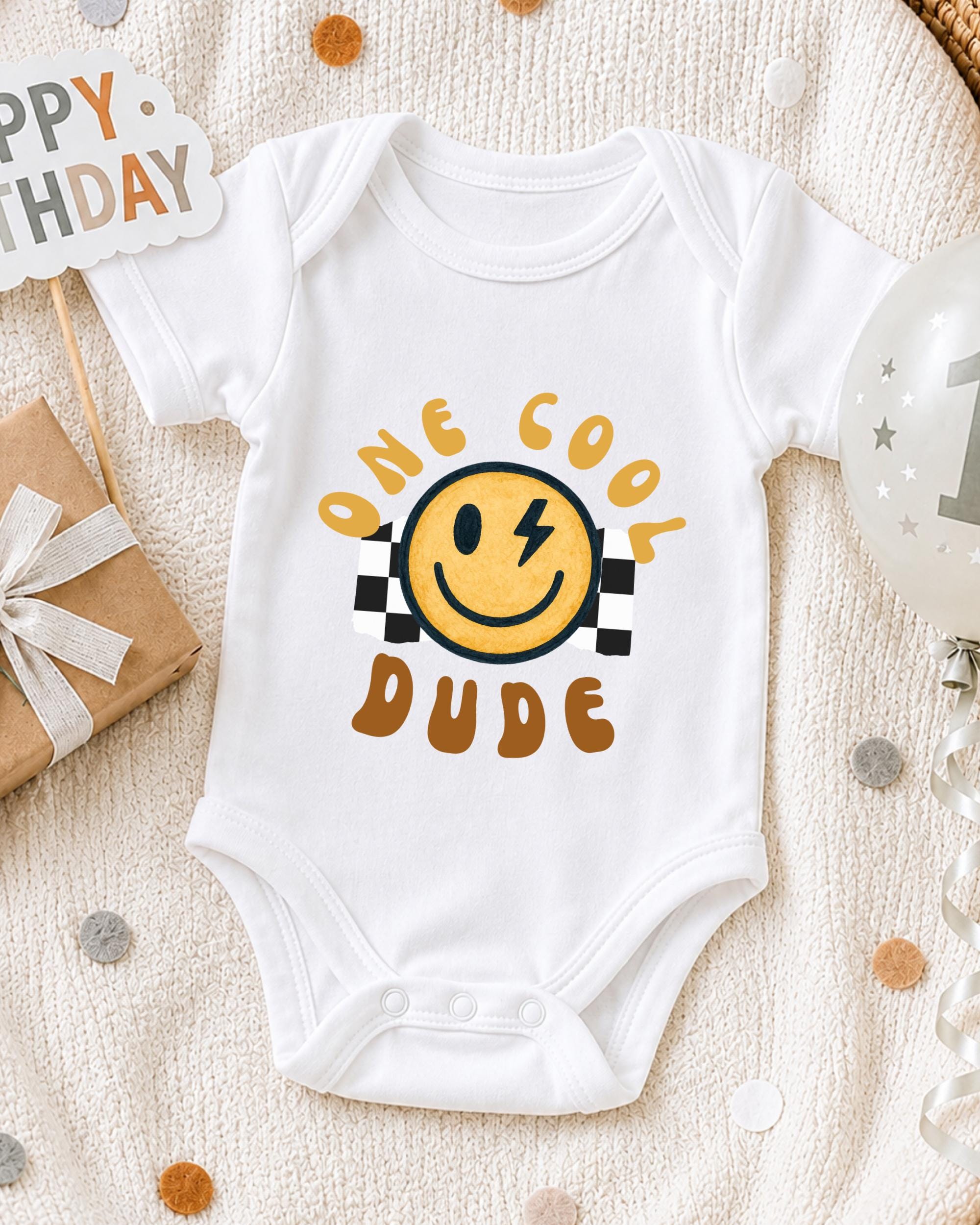 A white onesie with a yellow smiley face and the text "One cool dude" printed on it, along with a "Happy Birthday" sign and a birthday balloon.