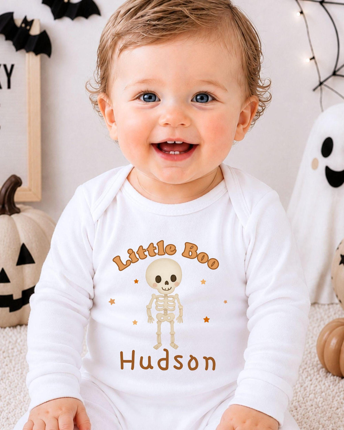 A smiling baby boy wearing a white onesie with a skeleton and the text "Little Boo Hudson" is sitting in front of Halloween decorations.