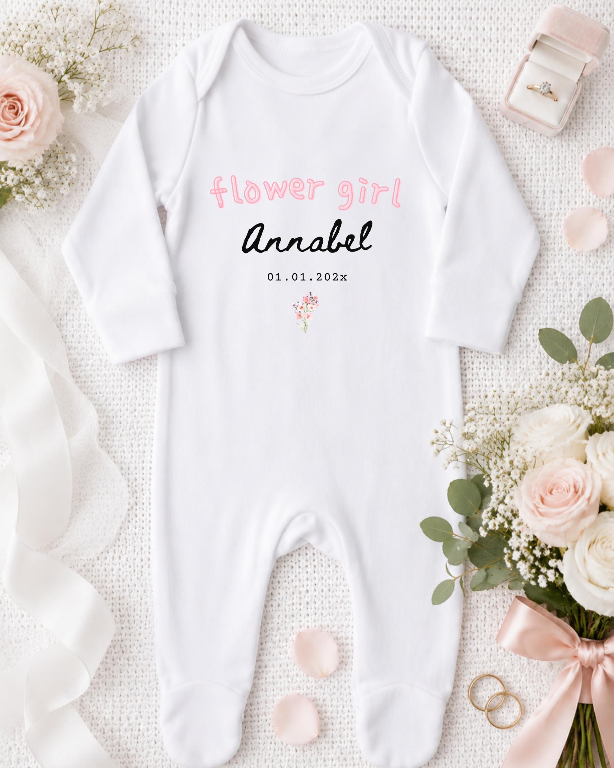 A white onesie with the text "flower girl Annabel" printed on it, along with a date "01.01.2023" and a small flower graphic. The onesie is placed on a white background, surrounded by pink flowers, a pink ribbon, and a pink box containing a ring.