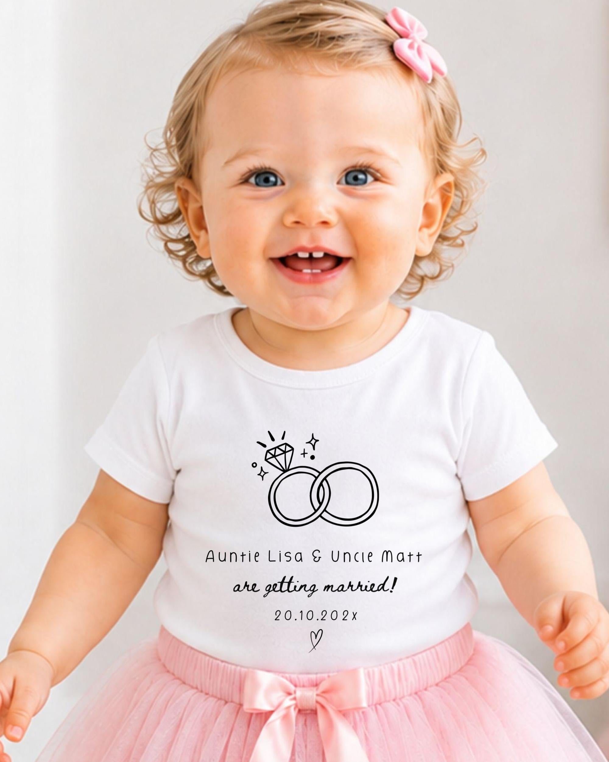 A smiling baby girl wearing a white t-shirt with a graphic of a ring and text that says "Auntie Lisa & Uncle Matt are getting married! 20.10.2021".