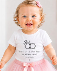A smiling baby girl wearing a white t-shirt with a graphic of a ring and text that says "Auntie Lisa & Uncle Matt are getting married! 20.10.2021".