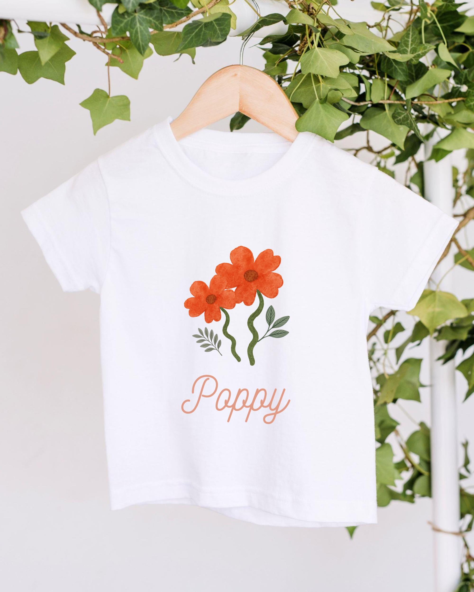 A white t-shirt with a floral design and the word "Poppy" printed on it, hanging on a wooden hanger.