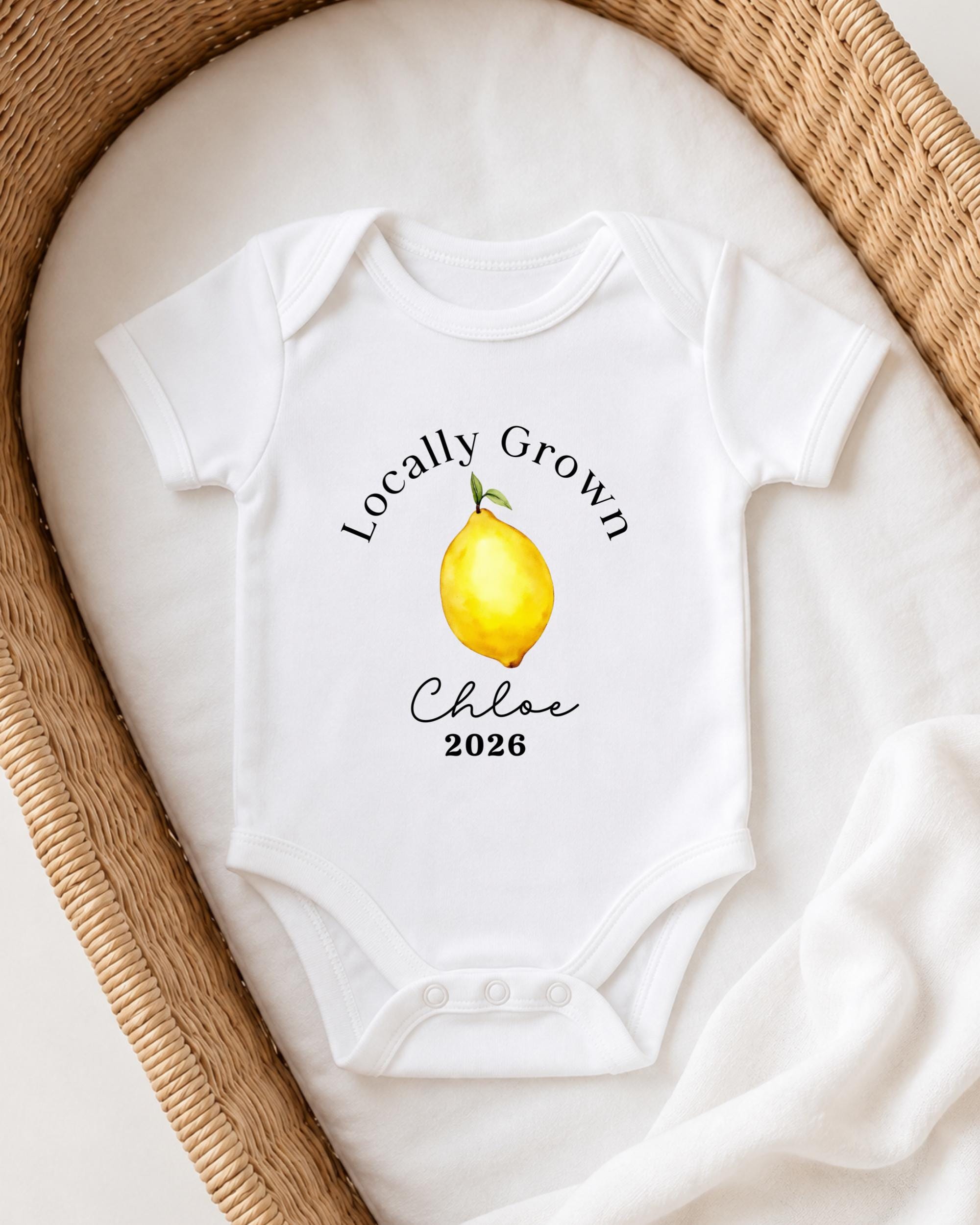 A white onesie with a lemon graphic and the text "Locally Grown Chloe 2026" printed on it, placed in a woven basket.