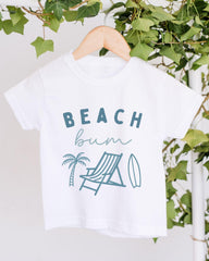 A white t-shirt with the text "BEACH butt" printed on it, along with illustrations of a beach chair, palm tree, and surfboard.